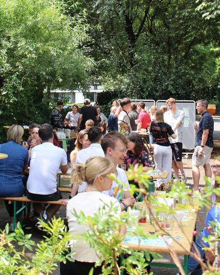 Eine sommerliche Freiluftveranstaltung mit vielen Menschen, die an Holzbänken unter Bäumen sitzen und sich unterhalten.
