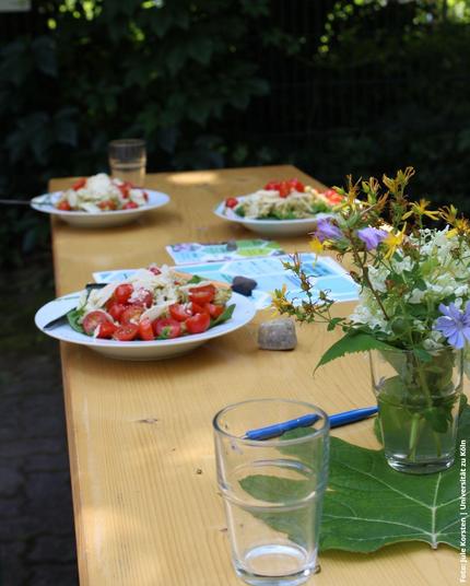 Ein Holztisch im Freien mit Teller mit Nudelsalat mit Tomaten und Basilikum, sowie ein Blumenstrauß in einem Glas.
