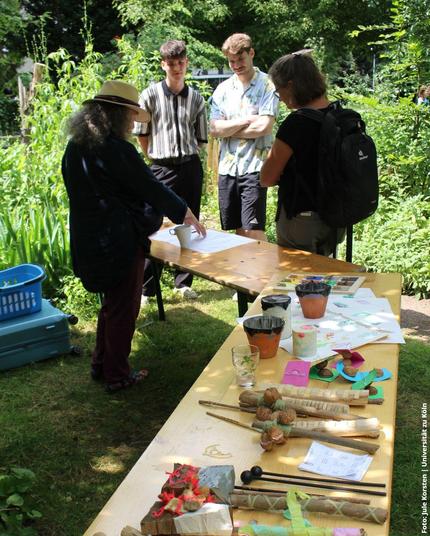 Eine kleine Gruppe steht im Garten vor einem Tisch, auf dem Bastelmaterialien und ein Blatt Papier ausgebreitet sind.