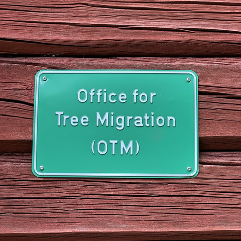 A green sign reading “Office for Tree Migration” posted on a red wood building wall