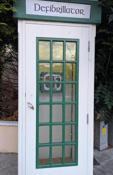 An old white and green Irish phone booth, with 'defibrillator' written in black letters on top, and an emergency semi automatic defibrillator hung inside