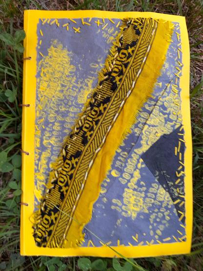 The cover of a yellow-blueish art book showing a diagonale of a silk strip on a maize cobble print. The book cover is made with sun-yellow cardboard. The cover itself: an abstract print collage of blue coloured krafft paper with a yellow print of maize cobbles and intuitive yellow embroidery at the borders. A stripe of yellow saree silk waste with a beautiful black pattern is sewed on the cardboard with white embroidery.