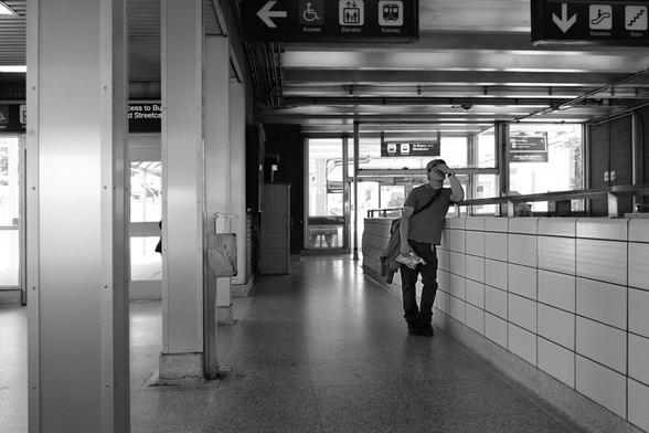 The main level of the station. In the background are the doors leading to the outdoor bus and streetcar platform. At the top of the frame are signs indicating the elevator, the escalators. At left are two of the pillars that hold up the ceiling, running from top to bottom of the frame. Just right of centre in the midground, a man leans against a tiled half wall, his hand over his eyes, maybe in fatigue or maybe from some deep emotion.