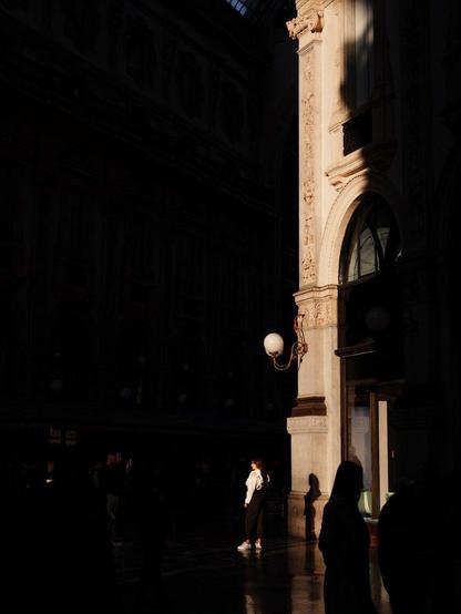 A young woman is visible against a very dark area as she's brightly lit by low sunlight. She is posing for a photograph and she projects a shadow against the wall behind her. A spherical lamp stands out above her, attached to an elegant wall. Silhouettes of other people are also visibile.