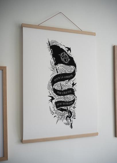 Photo d'une affiche accrochée au mur, qui représente un drapeau noir entouré de végétation, sur un fond blanc. Sur le drapeau on peut voir un logo anarchiste végétalisé ainsi que la mention de 3 phrases sur 3 étages : "pour la liberté", "par l'équité", et "avec entraide".