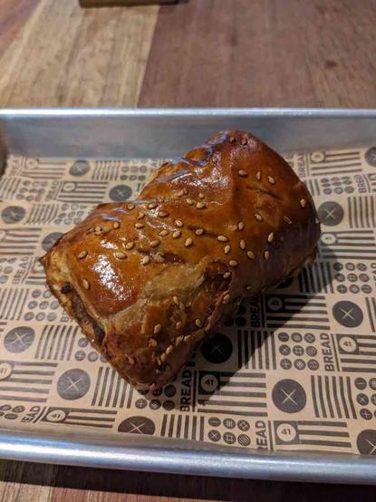 Portrait of a sausage roll on sheet of branded brown paper on an aluminum tray on a rustic wood table, the sausage roll is nicely browned with an egg glaze that holds a sprinkling of sesame seeds. One end of the sausage roll shows hints of the roasted sausage inside. It looks delicious.