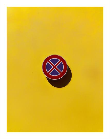 The photo is of a circular no-stopping (divieto di fermata) sign mounted on a bright yellow textured wall. The sign is mostly red with a blue central circle intersected by a red X with white trim. The sign casts a shadow on the wall on the bottom right hand side.