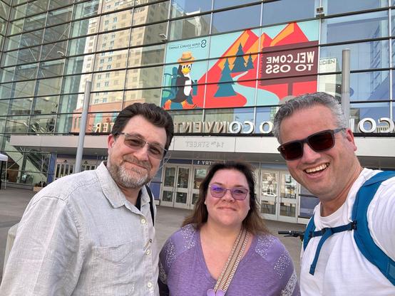 Karim Marucchi, Courtney Robertson, and Joost de Valk smiling outside the Colorado Convention Center in Denver. Behind them is a large glass facade featuring a colorful “Welcome to OSS” banner with a cartoon penguin wearing a hat and backpack, set against a mountain and forest backdrop. Karim is on the left wearing a light grey shirt, Courtney is in the center wearing a purple top and purple glasses, and Joost is on the right wearing a white shirt, sunglasses, and blue backpack straps.