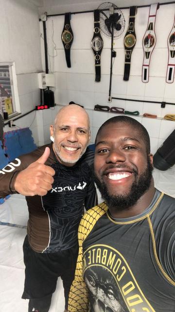 Dois homens negros sorrindo para a foto vestidos com roupas de treino de luta agarrada tendo ao fundo alguns cinturões de campeonatos de jiu-jitsu. Na esquerda sou (faixa-branca) eu e na direita meu professor e faixa-preta de jiu-jitsu.
