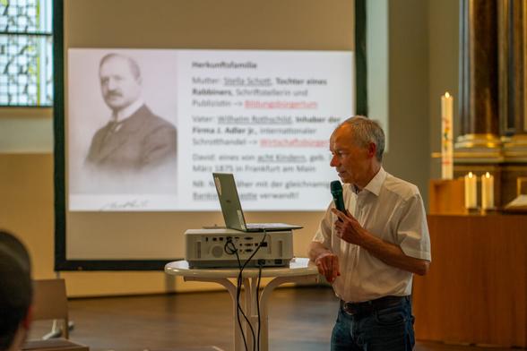 Dr. Markwart Herzog an einem Stehtisch, darauf ein Projektor und ein Laptop. Im Hintergrund ist das Bild erkennbar, das der Projektor "wirft", darauf in Schwarz-Weiß links ein Mann, rechts Schrift, nicht zu lesen. Rechts hinter Herzog im Anschnitt der Altar mit brennenden Kerzen. (Credit: Bernd Löser, Frankfurt am Main)