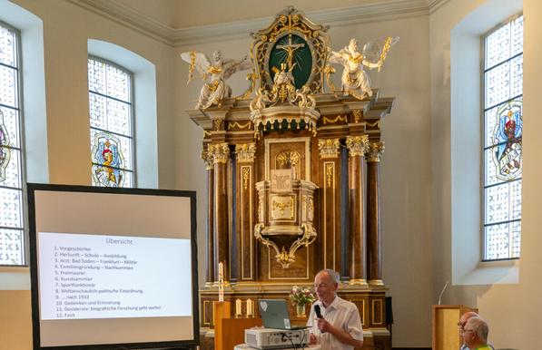 Ein prächtiger Altar gesäumt von Kirchenfenstern, davor eine Leinwand, auf der die Übersicht eines Vortrags zu lesen ist, mittig davor im Bild Markwart Herzog an einem Stehtisch mit Projektor und Laptop. (Credit: Bernd Löser, Frankfurt a. M.)