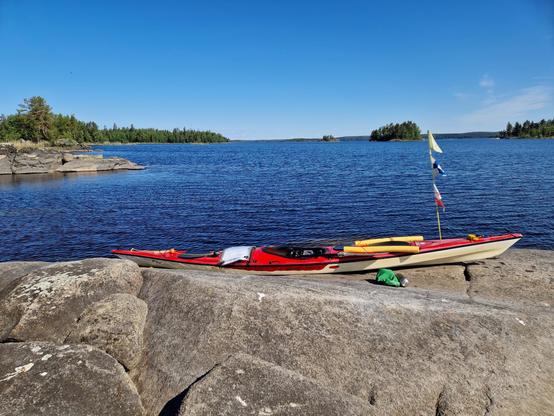 Matalalla rantakalliolla kajakki, vesitiivis eväspussi sekä termari. Kauempana lisää matalaa kalliota ja metsäisiä saaria. Taivas on lähes pilvetön.