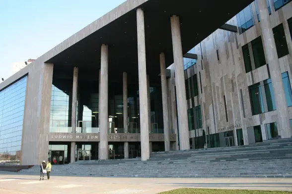 The image depicts a modern building that appears to be the Ludwig Museum, which features a contemporary architectural design. The structure has large glass windows and prominent columns, with a staircase leading to the entrance. There are also a couple of people standing in front of the building, and the setting appears to be a public space with some greenery.

Image Credits: Wikimedia / Peter Siroki from Budapest, Hungary / CC BY 2.0