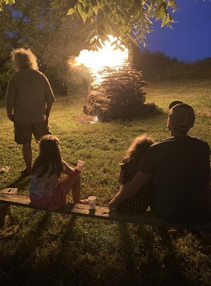 A large size bonfire, carefully constructed with crisscrossed layers of firewood, is just starting to burn.  A man stands a few yards away. On a bench are two girls and a father who are watching the fire.