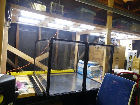 Homemade wooden shelving from 2x4s with overhead LED lighting holding a homemade 3-foot width by 3-foot deep by 2-foot tall box made from black 20x20mm extruded aluminum with 1/8th inch aluminum floor and 0.1-inch acrylic side and top panels with 2 closed swinging front doors.  A blue engineering height Steelcase engineering chair is sitting in front of the standing height desk durface.