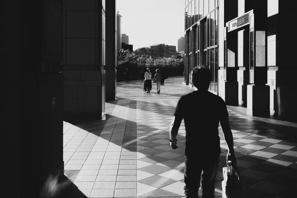 A black and white image featuring a person walking in the foreground with a bag, heading down a corridor with checkered flooring. In the background, three people are seen walking towards a bright outdoor space. Tall buildings create a stark contrast between light and shadow on the walls and floor.