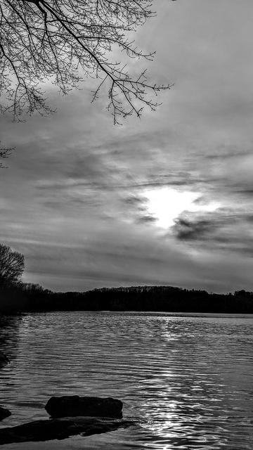 Monochrome image. Standing beside a large lake beneath a gray sky filled with dark clouds and thick haze. The lake is surrounded by a forest of still leafless hardwoods, the naked branches of a tall tree frame then top left of the pic. A pair of large, flat boulders partially submerged just offshore are casting long shadows towards us in the evening light. The rippled lake surface reflects the dark gray sky. The sun is almost burning through the clouds not far above the far shore, a bright spot in the gloom, its light creating a narrow trail of bright swirls on the waves.