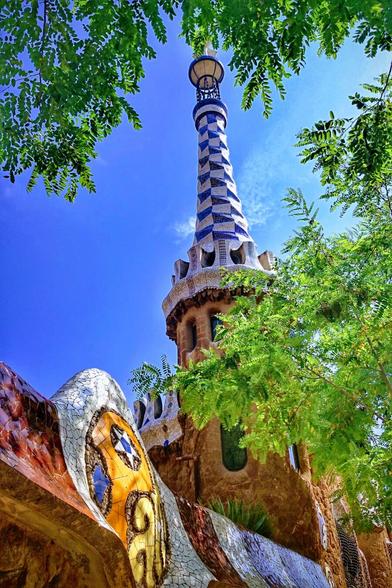 A low-angle shot of the colourful, mosaic-adorned architecture of Park Güell in Barcelona. A tall, checkerboard-patterned tower rises against a vibrant blue sky, framed by lush green tree leaves in the foreground, creating a sense of depth and natural beauty.
