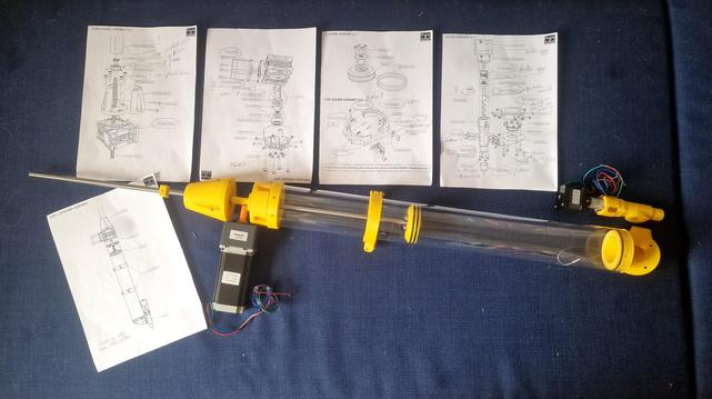 Delar till en blivande lerprinter utlagda på en mörkblå soffa: 

Fem A4-sidor med teknisk konstruktionsritning, en större motor med worm gearbox, en mindre stegmotor, ett 70 cm långt transparent rör och en lika lång gärngstång. 

Allt är delvis ihopsatt.