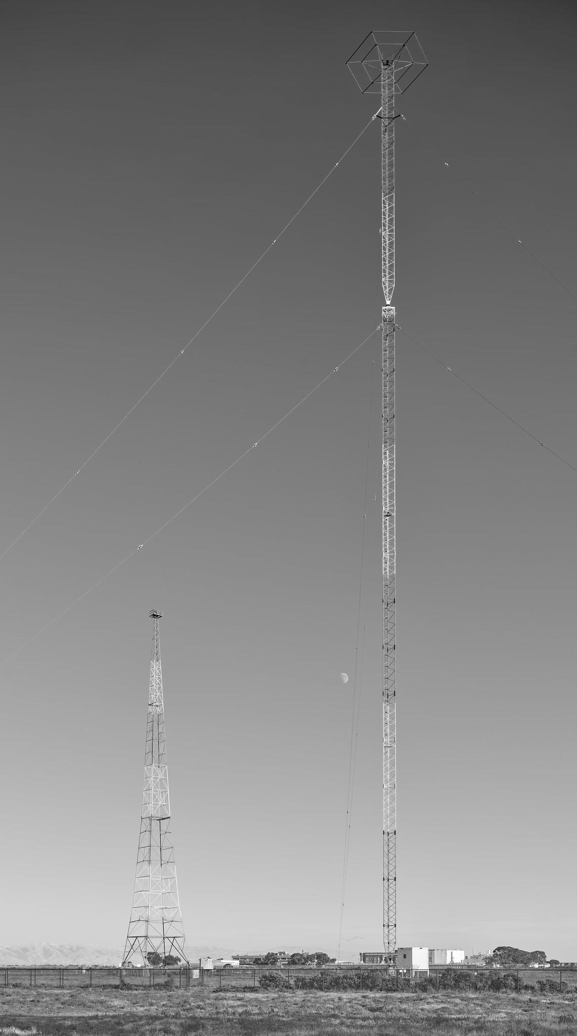 Two radio antenna masts in a field. The taller mast, on the right, is a guyed tower with a hexagonal structure at the top. The shorter mast on the left, toward the background, is freestanding.