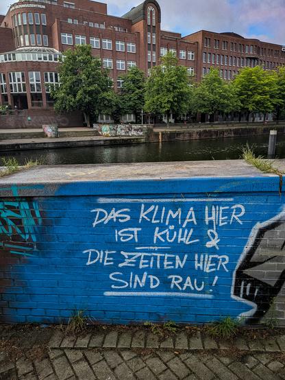 Eine Mauer am Mittelkanal die in weißer Schrift auf blau besprüht ist mit "Das Klima hier ist kühl & die Zeiten hier sind rau!"