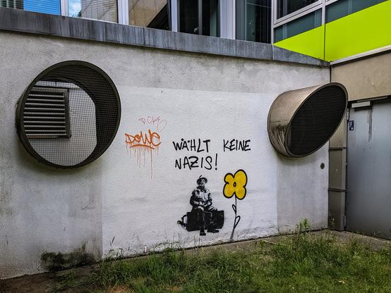 Eine weiß gestrichene Mauer mit Lüftungsrohren. Sie ist besprüht mit einer gelben Blume, einer schablonierten Person und dem Spruch "Wählt keine Nazis!"