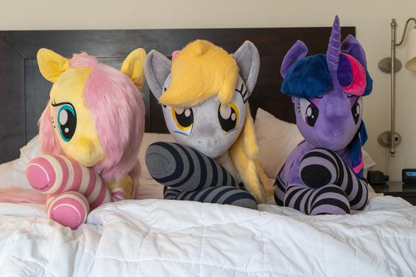 Three lifesize pony plushies, Fluttershy, Derpy, and Twilight. They lie comfortably on a bed with their front legs crossed.