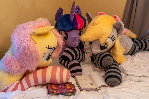 Three lifesize pony plushies, Fluttershy, Twilight, and Derpy. They lie on a bed with a few scattered books staged as if they were reading together.