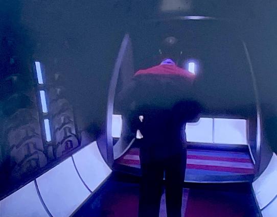 A photo of a television, showing a man in a black uniform with a red band across the shoulders walking away down a corridor. At the junction with another corridor the floor has the blue-pink-white-pink-blue stripes of the trans flag.