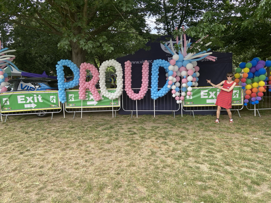 A photo taken by a “Proud” sign made of balloons and in the Trans flag colour. I’m to the right in a warm autumn dress, with a red bell choker and 3 pride pins, posing with both my hands pointing at the sign.