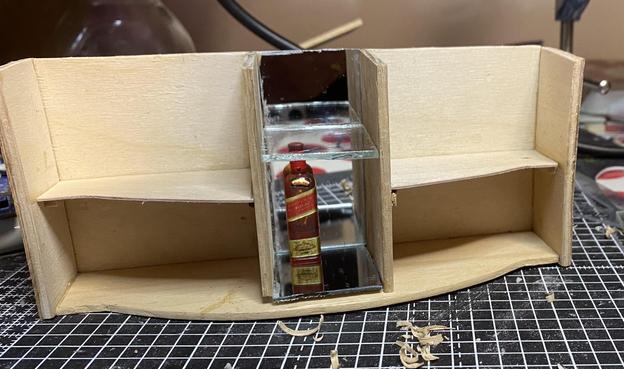 The bare carcass of a dollhouse sideboard with a bottle of scotch in the drinks cabinet for scale. The carcass is of light modelling wood obeche and plywood and the mirrored section is in the middle. There is no top and no doors or feet.