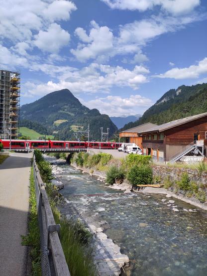 Foto. Ein Bach mit klarem Wasser und ein roter Zug zwischen Häusern in Berglandschaft.
