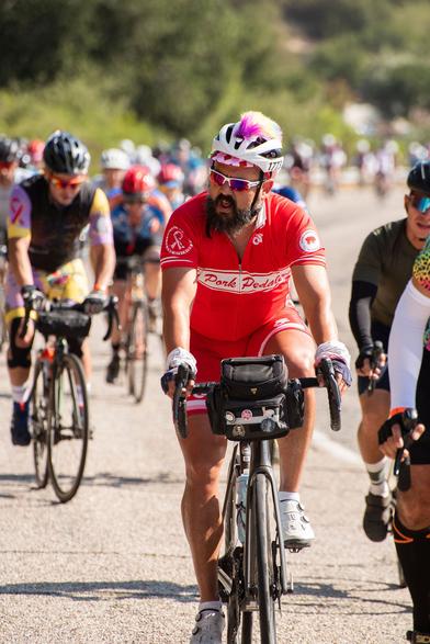 Jorge riding his bicyle wearing an all red cycling kit that says "Pork Pedalers" on the front. He has a white helmet on that has a rainbow mohawk on top.