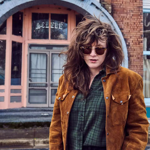 A white woman with long, flowing black hair holds her hands in pants pocket. She wears a brown suede jacket, green flannel, and horn-rimmed aviators. She stands in front of a rounded doorway.