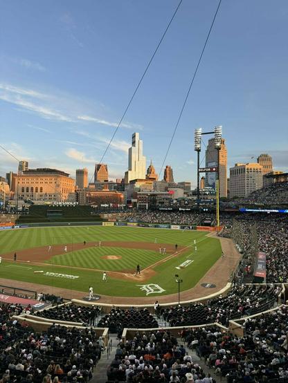 A baseball game is taking place in a packed stadium with a city skyline visible in the background under a clear sky.
