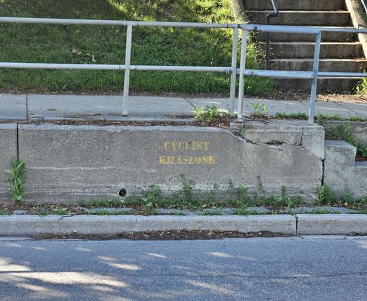 There is a concrete wall next to a road without a shoulder.  Spray painted in yellow block letters, it says "CYCLIST KILL-ZONE" .