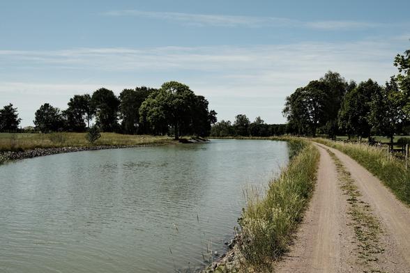 A serene landscape featuring a a curved canal, Göta Canal, bordered by grassy banks and trees, with a dirt path running alongside the water. The sky is clear with a few clouds.