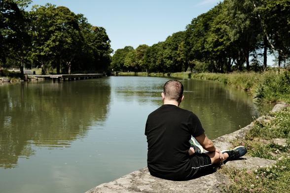 A person sits on a stone by a calm canal, facing the water surrounded by lush greenery and trees. The scene is tranquil, reflecting a sunny day with clear skies.