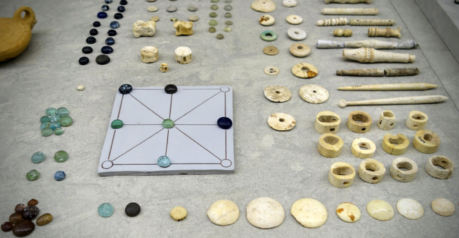 In einem Museum sind archäologische Funde ausgestellt. Im Foto zu sehen: Spielsachen wie eine Art Mühlespiel mit kleinen Steinchen oder pfeifen- bzw. flötenähnliche Kleinigkeiten.