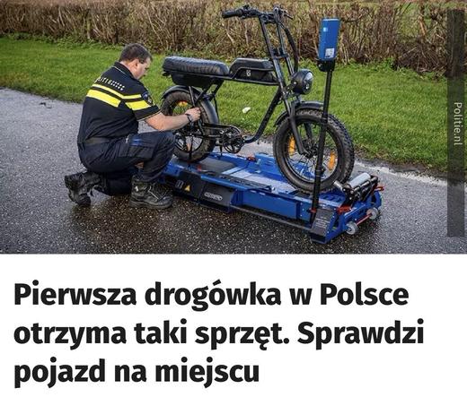 Policjant klęczy obok roweru elektrycznego na niebieskiej platformie do badań. Scena rozgrywa się na zewnętrznej drodze, z trawą w tle. Obraz sugeruje wprowadzenie nowego narzędzia do kontroli pojazdów dla policji w Polsce.