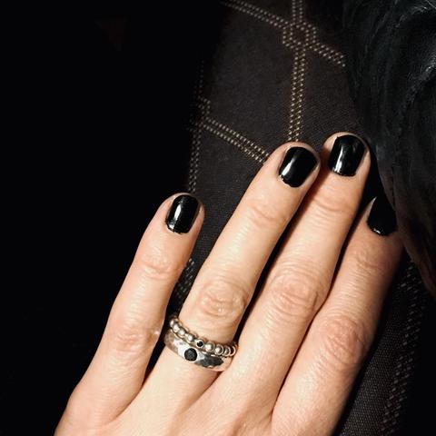 A hand with black nail polish is shown, wearing a textured silver ring with a black stone. The background features dark, patterned stockings.
