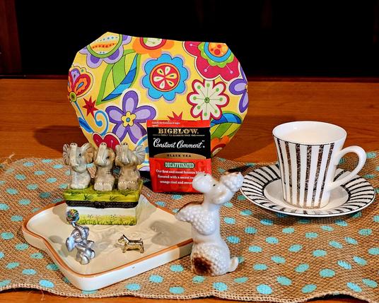 Bright tea tableau on turquoise- polkadotted burlap with a vivid tea cozy print of psychedelic flowers and Constant Comment black tea from Bigelow. There is a boldly platinum- striped demitasse set, and a ceramic tray shaped like a slice of white bread, upon which are a trio of elephants with trunks raised in unison opposite a pewter elephant balancing a rainbow crystal ball on its snout, about to toss it to a circus poodle while a tiny terrier looks on.