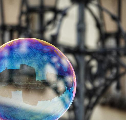 Das Bild zeigt eine große, farbenfrohe Seifenblase, die vor einem verschwommenen Hintergrund mit einem kunstvollen Metallgestänge steht. Die Seifenblase reflektiert ein buntes Muster aus Blau, Violett und Rosa, das durch die Lichtinteraktion entsteht. Innerhalb der Blase ist ein Spiegelbild einer Stadtlandschaft zu sehen, die sich in der glatten Oberfläche der Blase widerspiegelt. Die Gebäude sind dunkel und kontrastieren mit dem leuchtenden Hintergrund der Blase. Der Hintergrund ist unscharf, was die Blase im Vordergrund hervorhebt.
