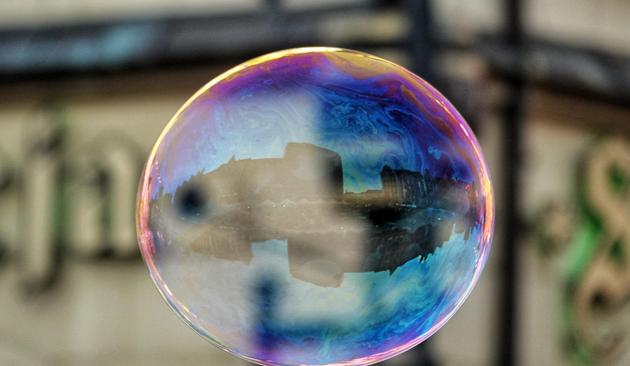 Das Bild zeigt eine große, glänzende Seifenblase, die in der Luft schwebt. Die Blase reflektiert die Umgebung und zeigt ein farbenfrohes Muster aus Blau, Violett und Gelb. Im Inneren der Blase ist ein Spiegelbild eines Gebäudes zu sehen, das sich in der Mitte der Blase befindet. Der Hintergrund ist unscharf und zeigt ein Gebäude mit einem Teil eines Schildes, das teilweise sichtbar ist. Die Blase ist die Hauptfokuspunkt des Bildes und fängt die Lichtreflexionen ein, was ihr ein lebendiges und farbenfrohes Aussehen verleiht.
―