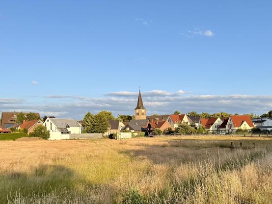 Blick auf ein kleines westfälisches Dorf mit roten und weißen Häusern, im Hintergrund eine Kirche mit spitzem Turm. Im Vordergrund eine goldene Wiese und ein Radweg unter blauem Himmel.