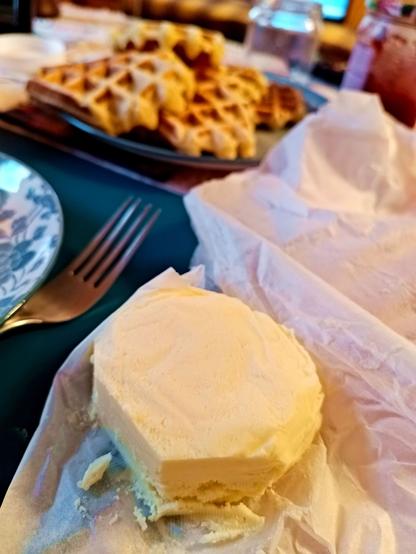 A round shaped piece of selfmade butter wrapped in grease paper. It's had pieces cut off on two sides. Behind it, a pile of fresh Belgian waffles on a plate and a fork.