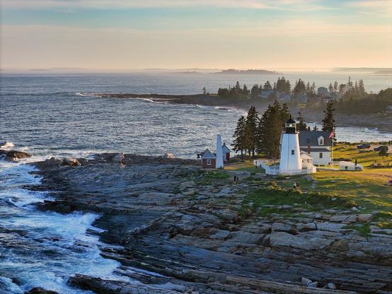 Pemequid Point Light sits on a rocky ledge in the evening sunlight. In the distance, rocky islands can be seen enshrouded in a bit of mist