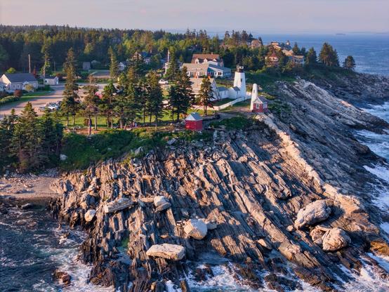 A lighthouse sits on a rocky prominence, while water crashes at it's base.
