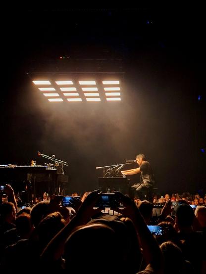 Trent Reznor, singer of Nine Inch Nails sits at a piano and sings on a small, low stage in the middle of the crowd with only one set of lights lighting up the completely dark concert arena