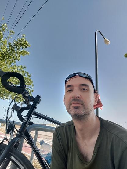 Yo de torso con un pañuelo en la cabeza (un paliacate) con gafas de sol encima. Mi bici negra sale detrás. Está entre medio de un aparcabicis. El cielo es azul y hay una farola detrás.